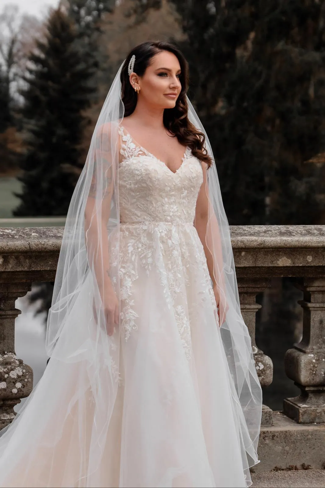 Braut mit Schleier und filigranem Spitzenkleid vor einer Steinbalustrade, gestylt mit natürlichem Braut-Make-up und weichen Wellen.