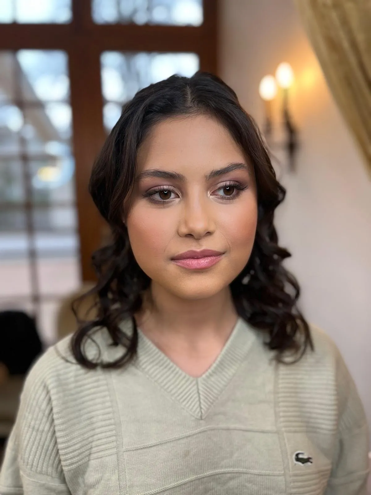Porträt einer jungen Frau in einem hellen Pullover, gestylt mit sanftem Augen-Make-up und offenen Locken in einem Wohninterieur.