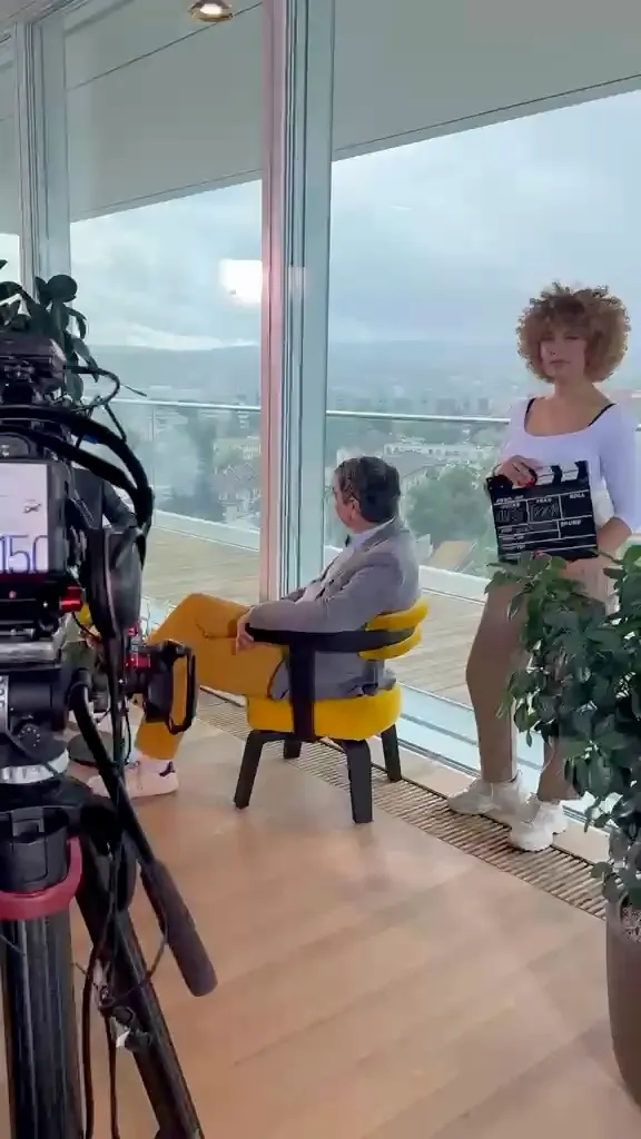 Filmset mit Kamera im Vordergrund — Sandra hält die Klappe bereit, während im Hintergrund eine Interviewsituation vorbereitet wird.